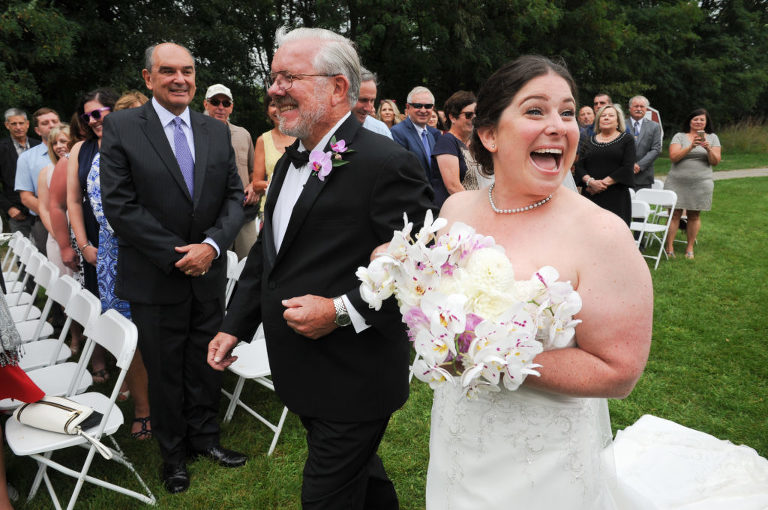 The bride and her father walk down the path to the ceremony site at the Indian Springs Metropark wedding in White Lake, Michigan as the bride spots someone to say "hi" to