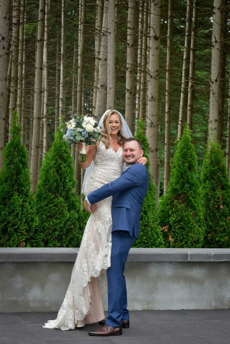 Using the dramatic lodge pole pines as a backdrop for wedding photos the groom lifts the bride off a ledge at Shepherd's Hollow's new wedding venue in Clarkston, Michigan.