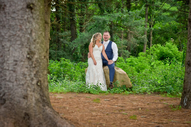 Sitting on a rock (got tired of climbing on stuff in her mega heels), the bride and groom pose for a photo in the woods at a wedding at Shepherd's Hollows Golf Club Clarkston, Michigan.