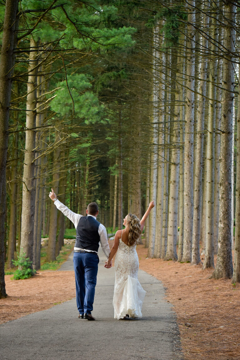 The bride and groom walk back to their wedding reception with their signature wave in amongst the lodge pole pines at a wedding at Shepherd's Hollows Golf Club Clarkston, Michigan.