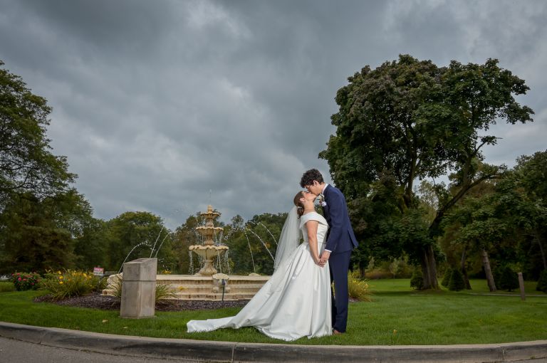 The bride and groom pose for photos at the fountain during their fall wedding at Addison Oaks Buhl Estates in Leonard, Michigan.